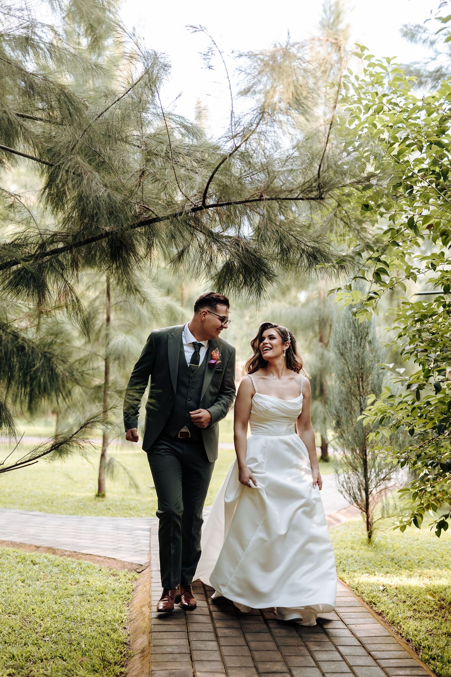 A romantic wedding portrait on a garden pathway with towering pine trees creating a natural backdrop.
