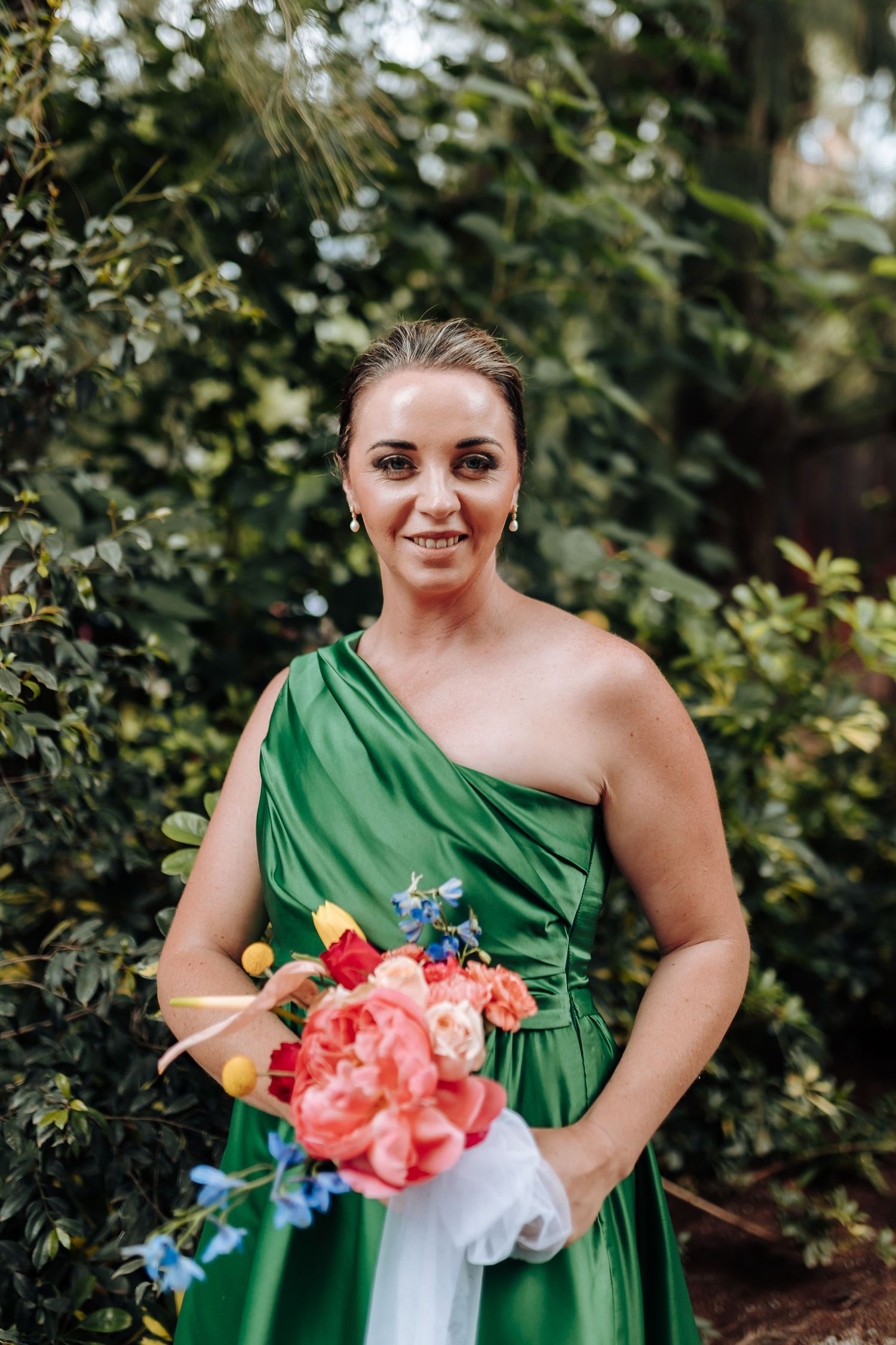 A bridesmaid in an emerald green one-shoulder dress holds a vibrant coral and white bouquet outdoors.