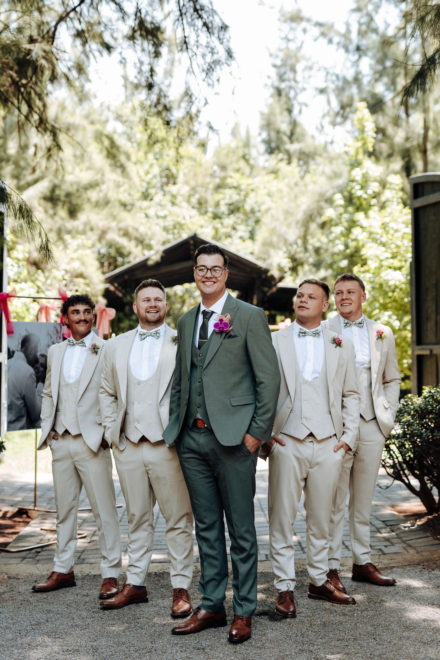 A wedding party group photo with groomsmen in light colored suits and one in dark green standing together outdoors.