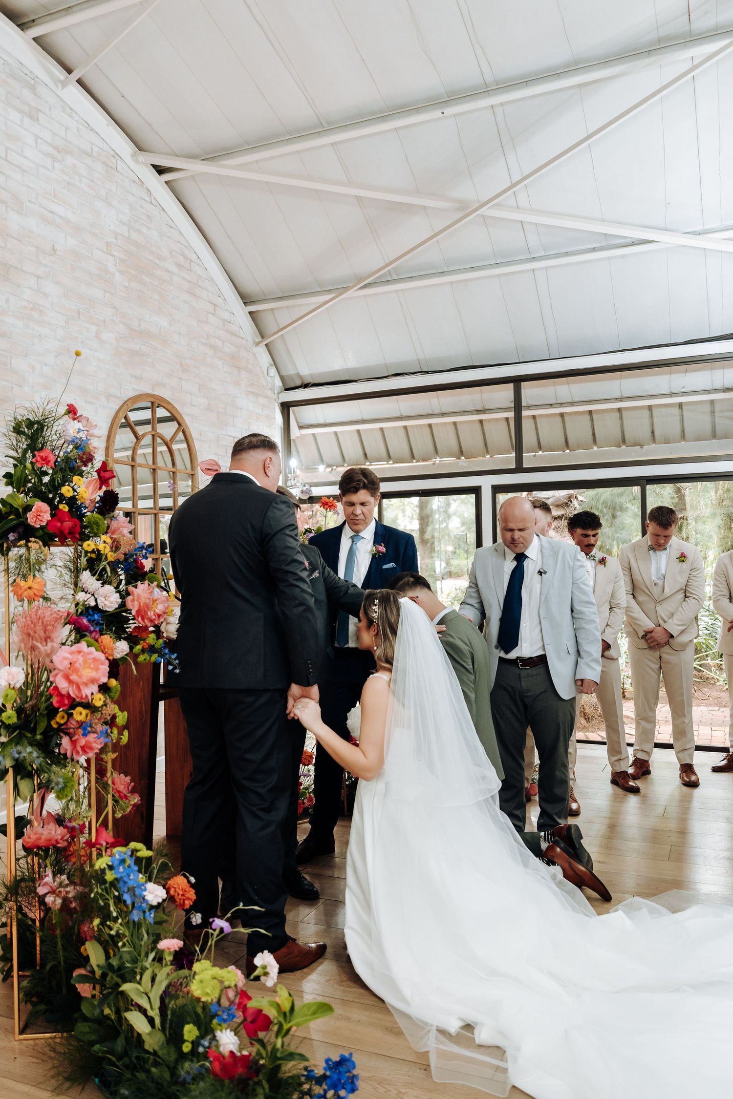 Wedding celebration in a white tent-style venue with colorful floral arrangements and guests mingling.