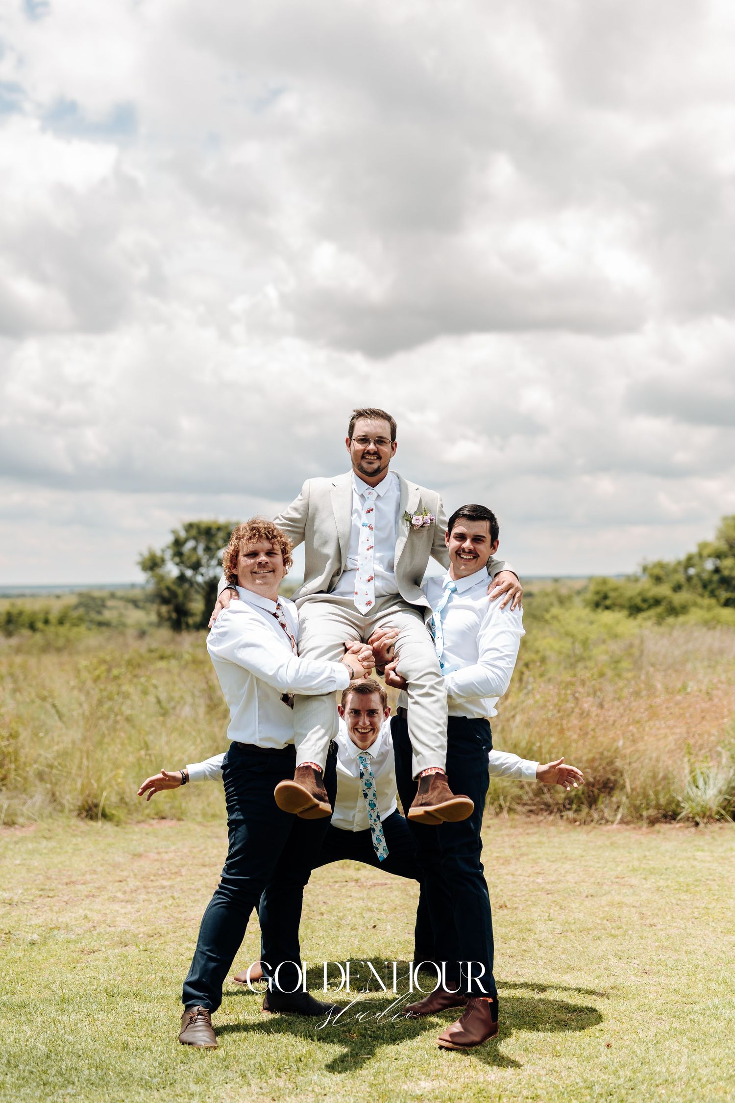 Groomsmen in white and black attire strike playful pose lifting one person in outdoor setting with cloudy sky.