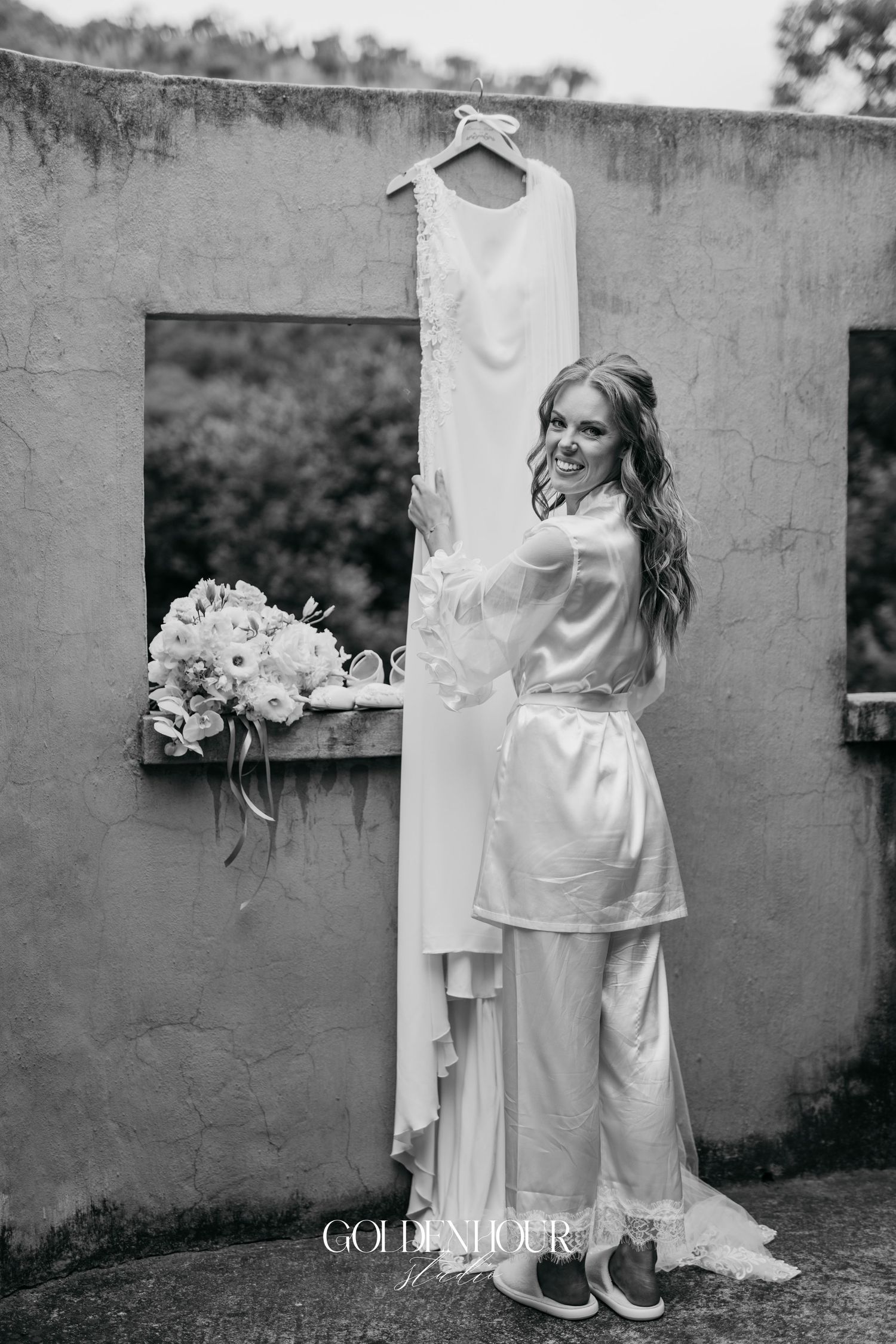 Black and white wedding photo showing elegant white dress details and floral bouquet against a textured wall.