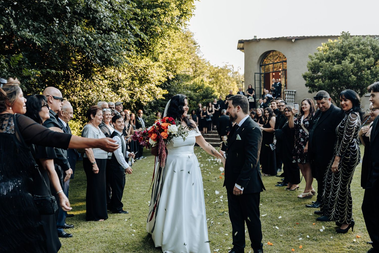 Wedding party and guests line up outdoors for a celebratory moment on a sunny day.