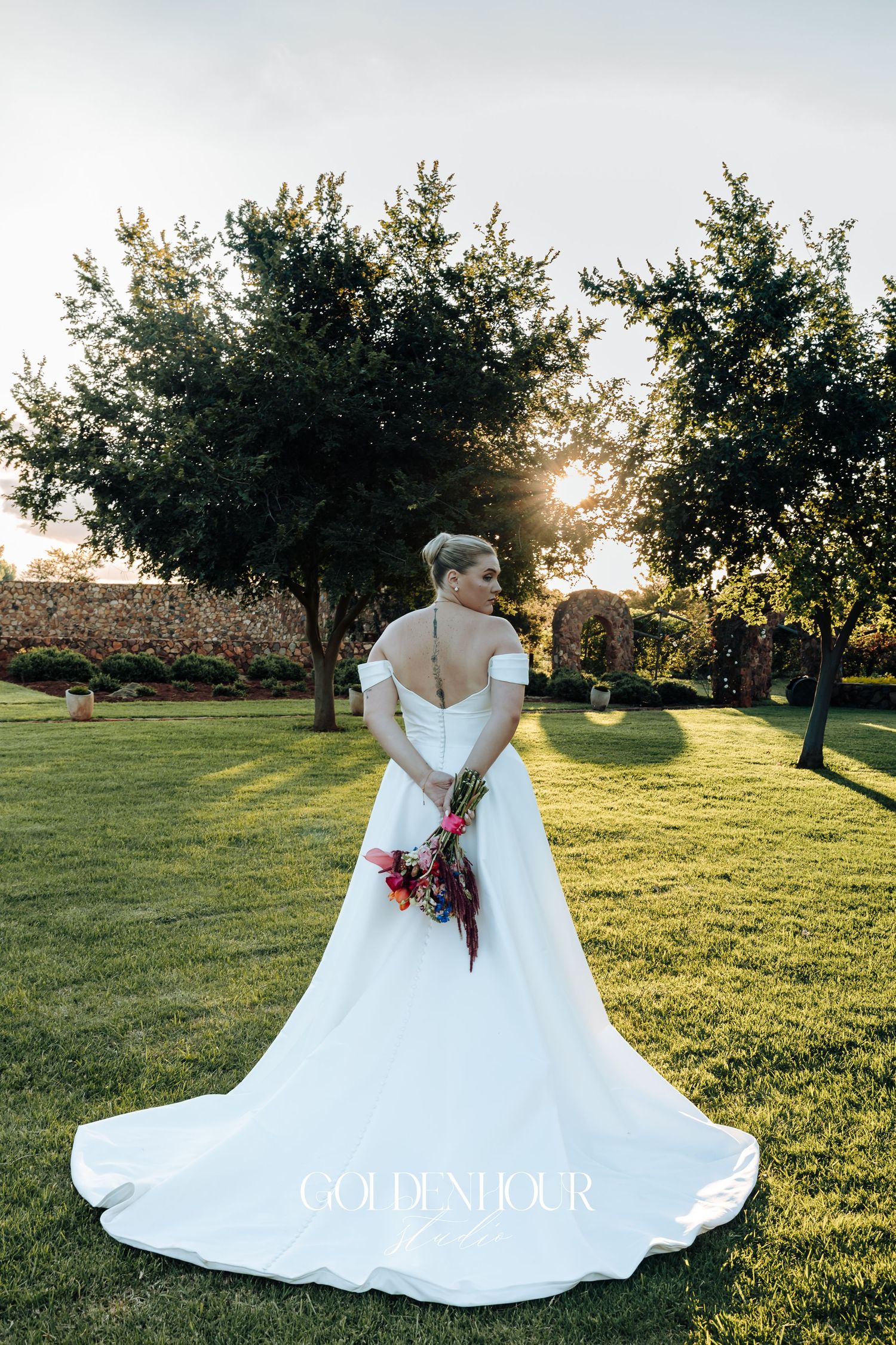 A bride in a flowing white wedding gown poses in a sunlit orchard during golden hour.