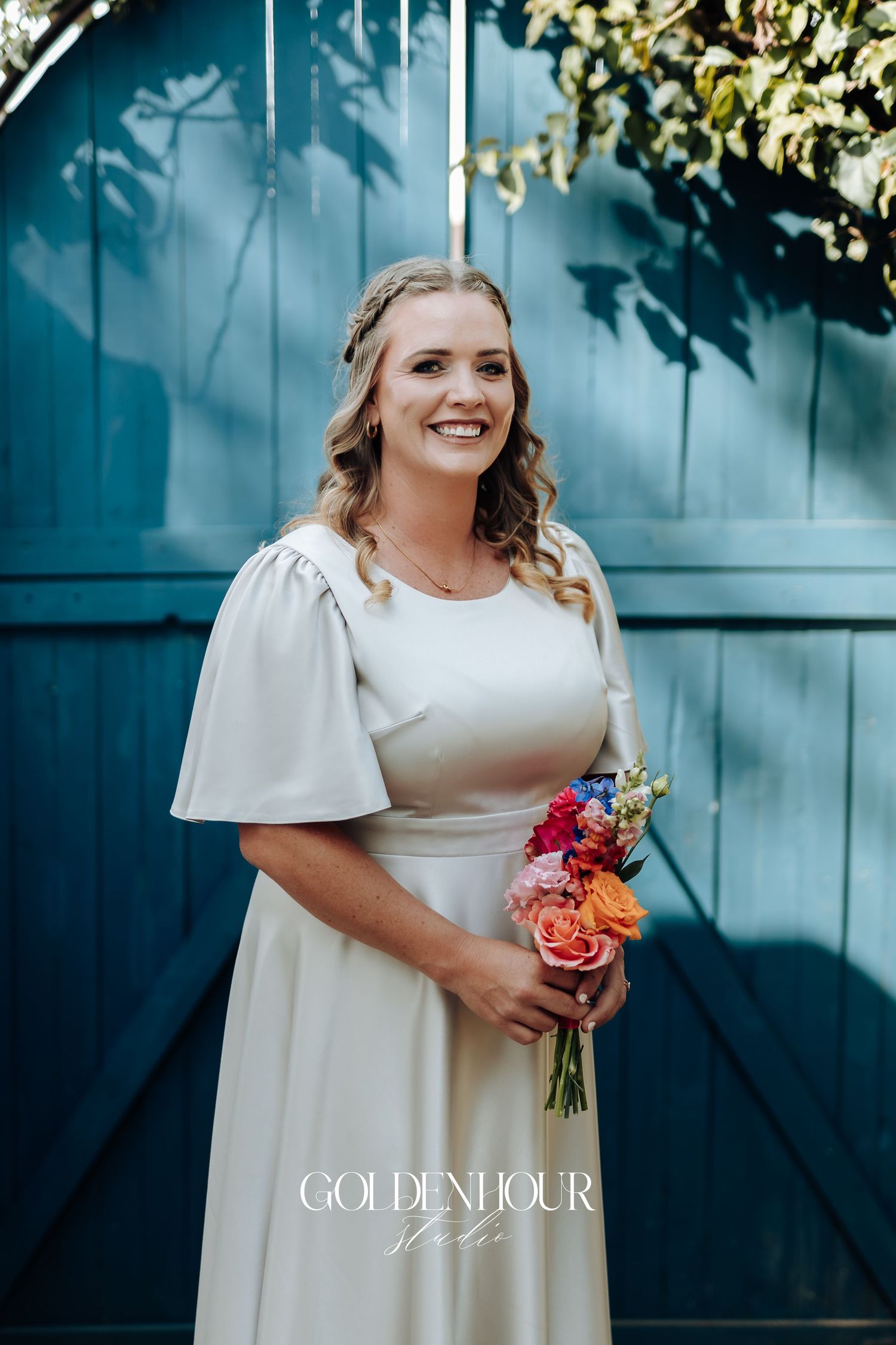 A bride in a classic white dress holds a vibrant coral and blue floral bouquet against a teal backdrop.
