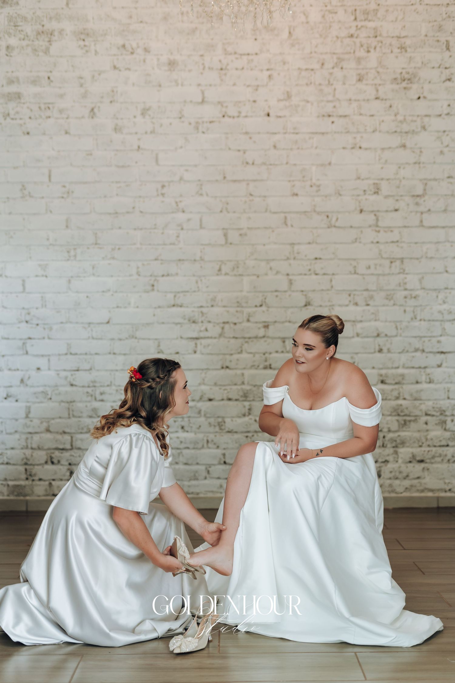 Two people in white dresses sit together on the floor against a brick wall, sharing a tender moment.