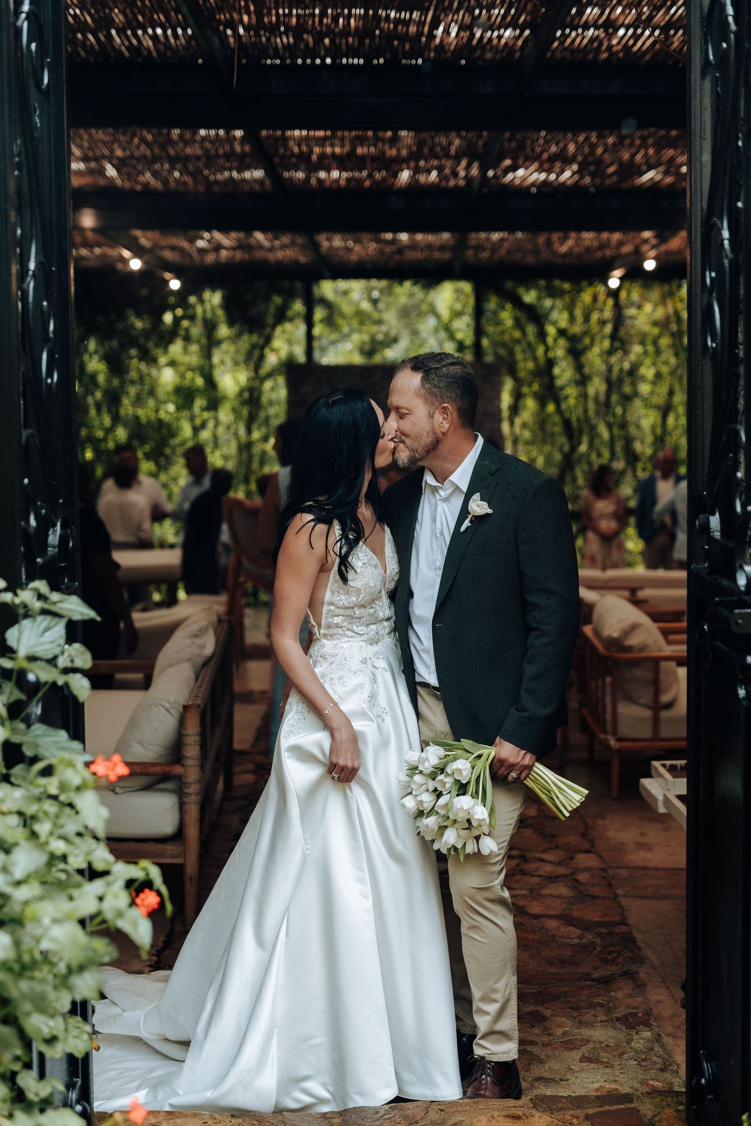 Bride and groom share a kiss at outdoor wedding venue surrounded by lush greenery, bride holding white bouquet.