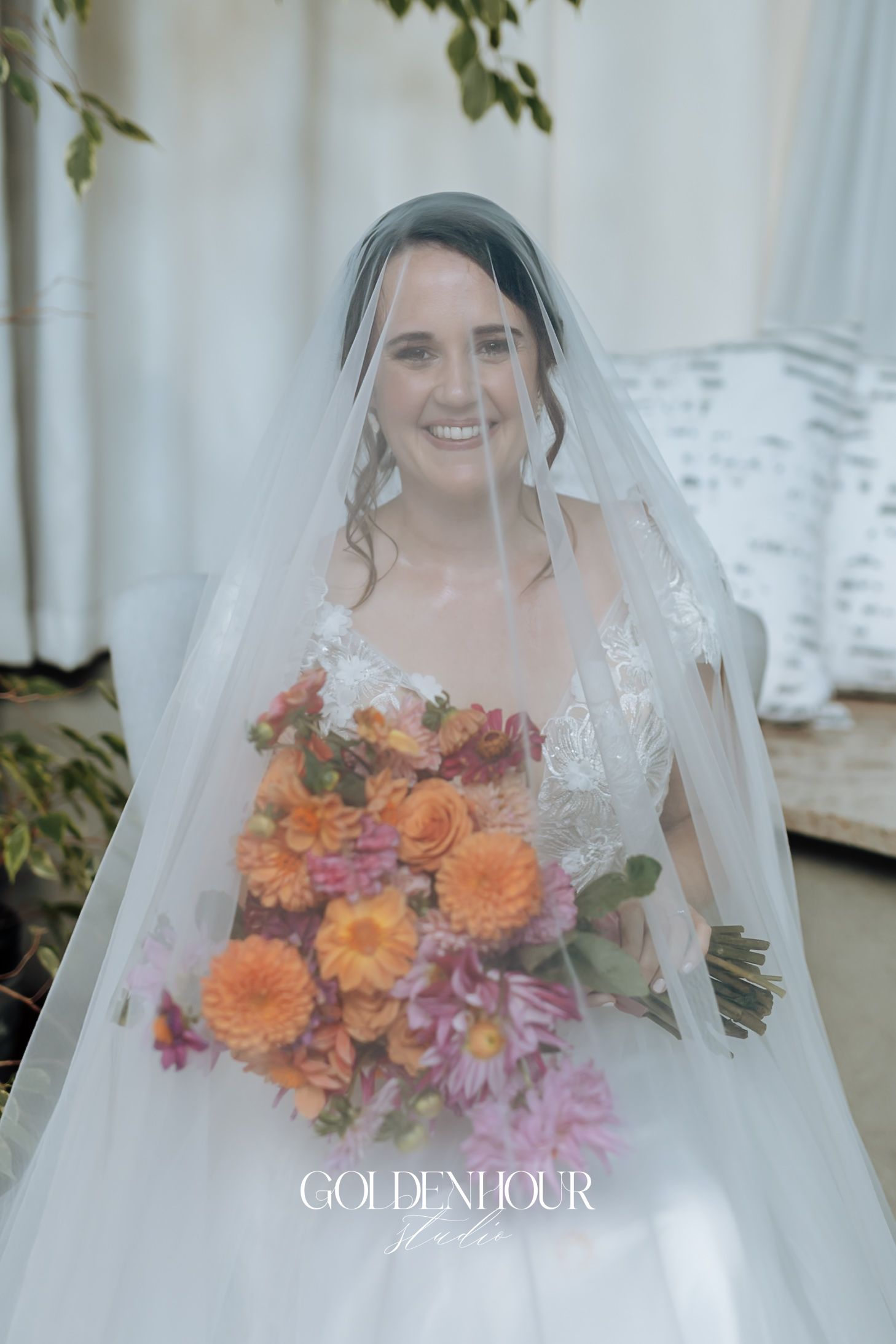 Smiling bride holding vibrant orange and pink bouquet under flowing white wedding veil.