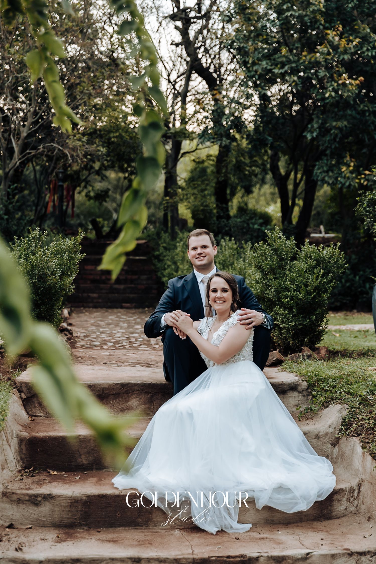 Smiling groom holds bride in white gown on rustic stone steps in a lush garden setting.