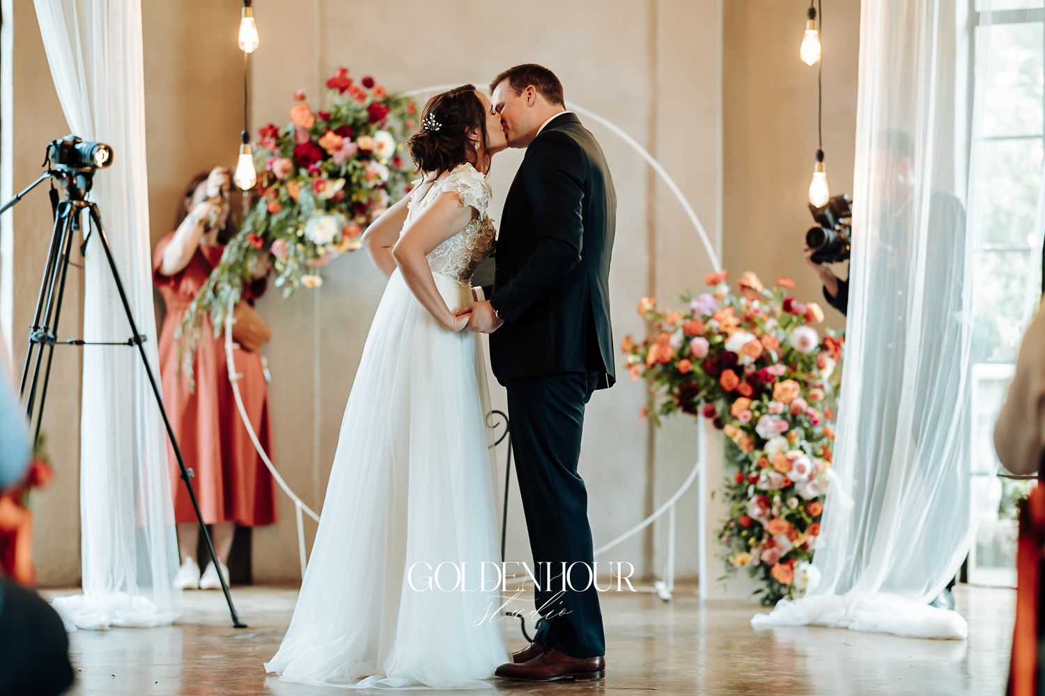 Bride and groom sharing first kiss at altar with colorful floral backdrop.