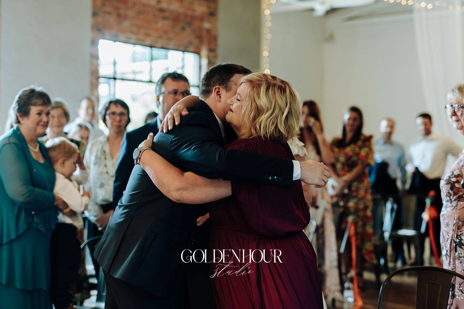 Two wedding guests share a heartfelt hug during an indoor wedding ceremony surrounded by seated guests.
