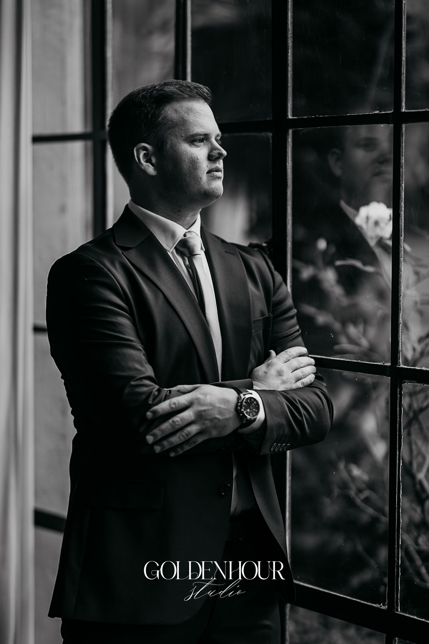 Dapper groom in a dark suit and tie gazing through a window, elegant black and white wedding portrait.