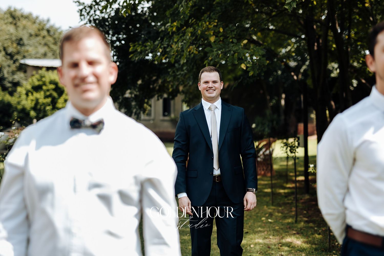 Groom in navy suit smiling at groomsmen dressed in white shirts with bow ties outdoors.
