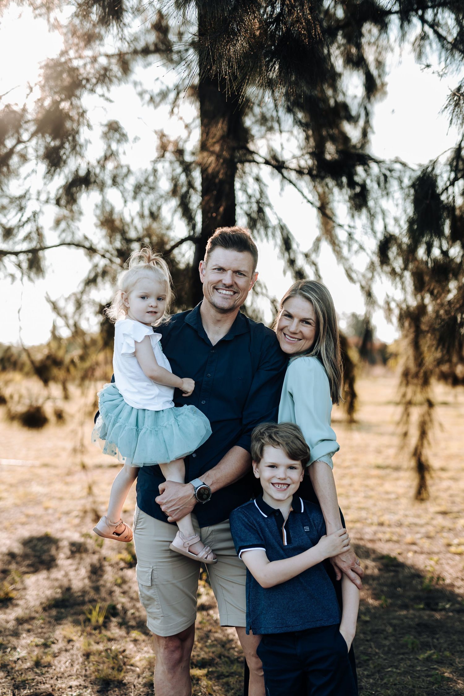 A family poses together outdoors on a sunny day with pine trees in the background.
