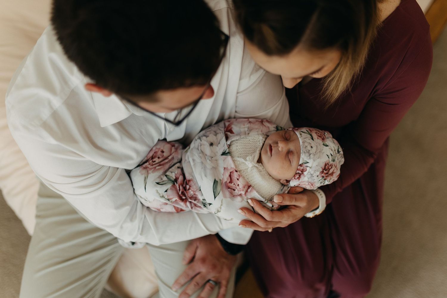 Newborn baby wrapped in floral swaddle blanket being held by parents in burgundy and white tops.