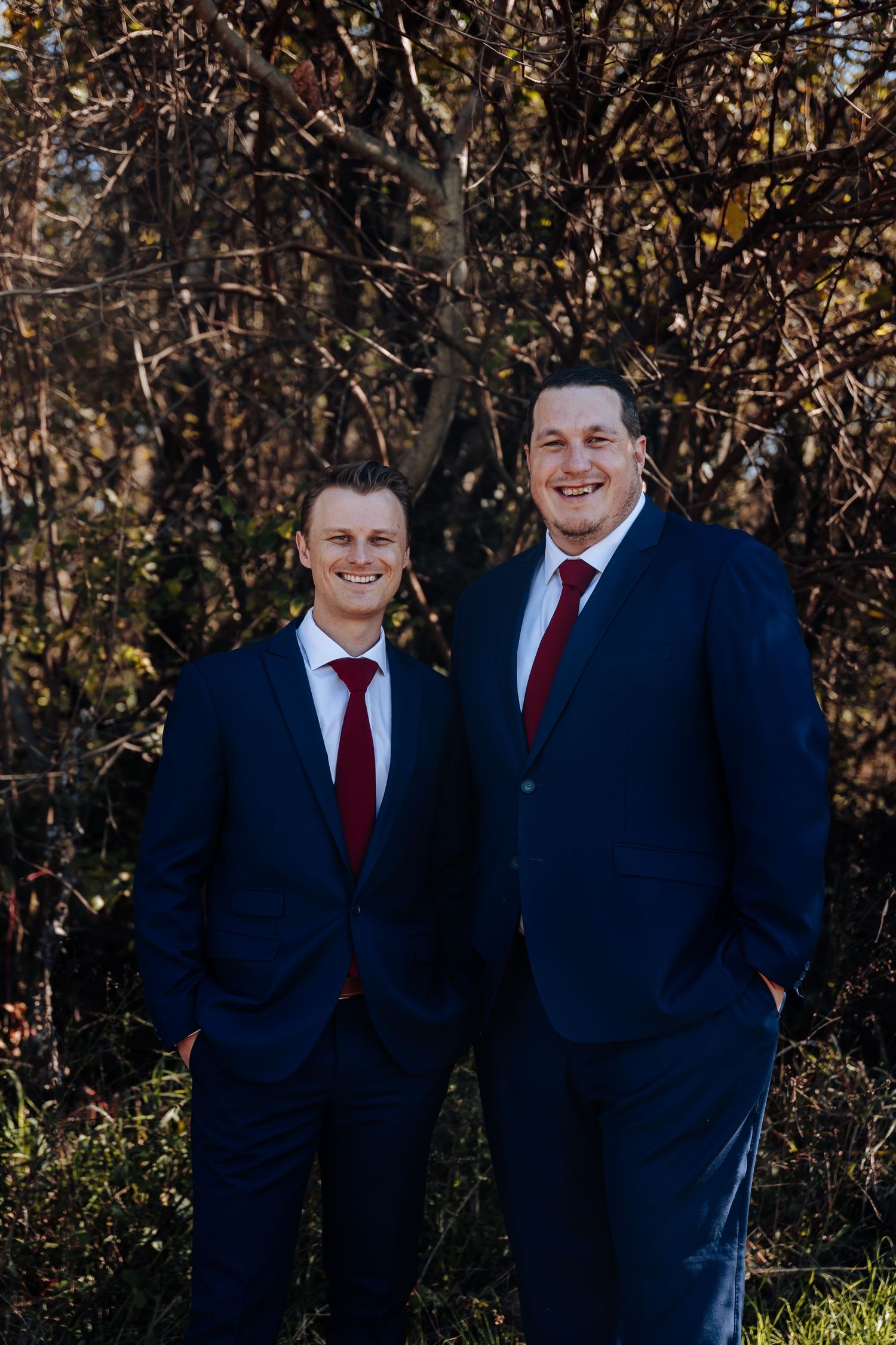 Two men in navy blue suits and red ties smiling outdoors in front of autumn trees.