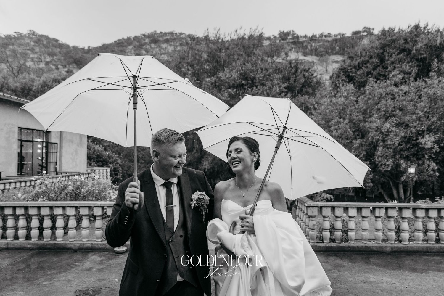 Black and white photo of laughing bride and groom sharing umbrellas in the rain outdoors.