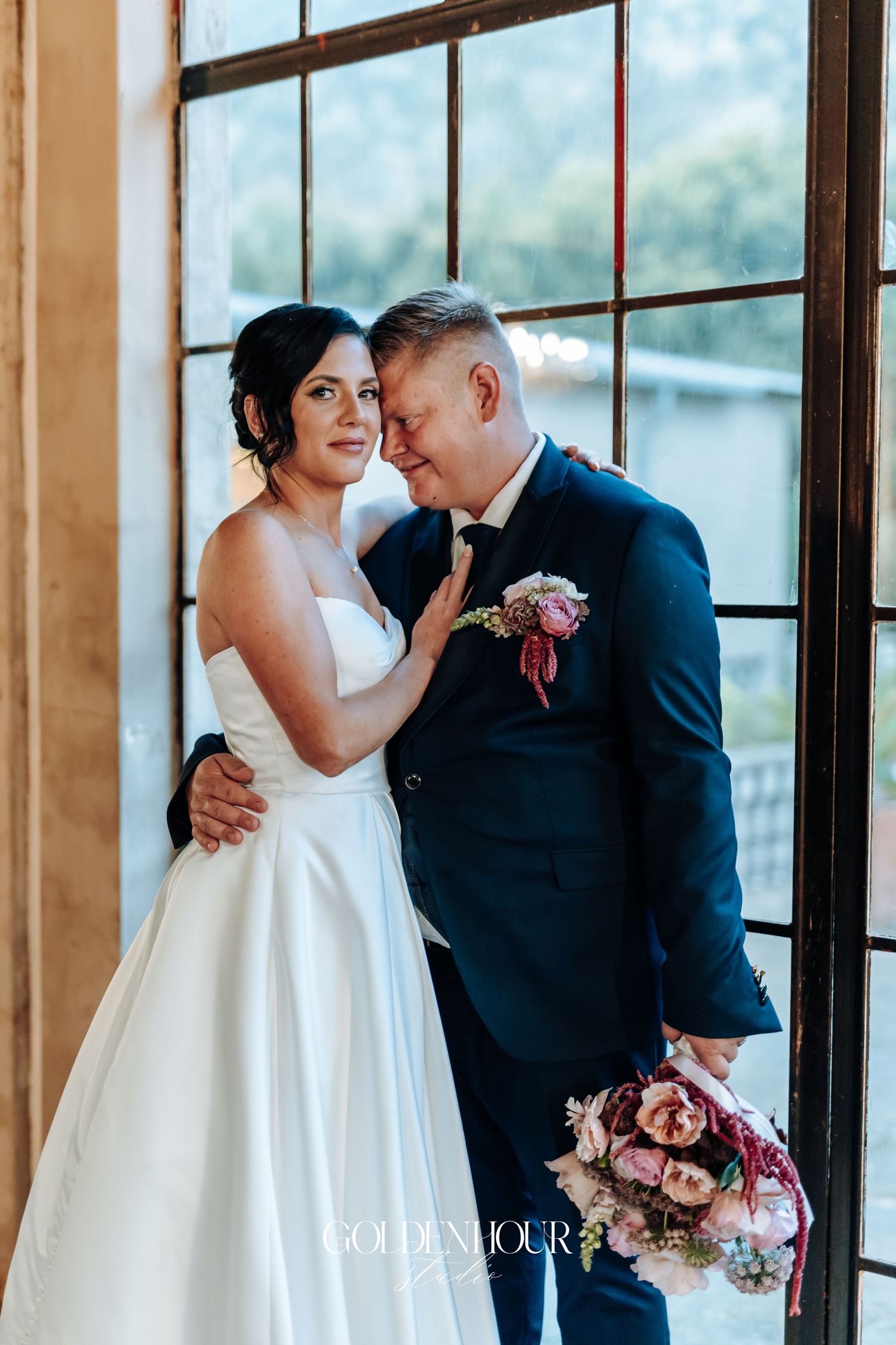 Bride and groom share tender moment by large window, she in white gown, he in navy suit.