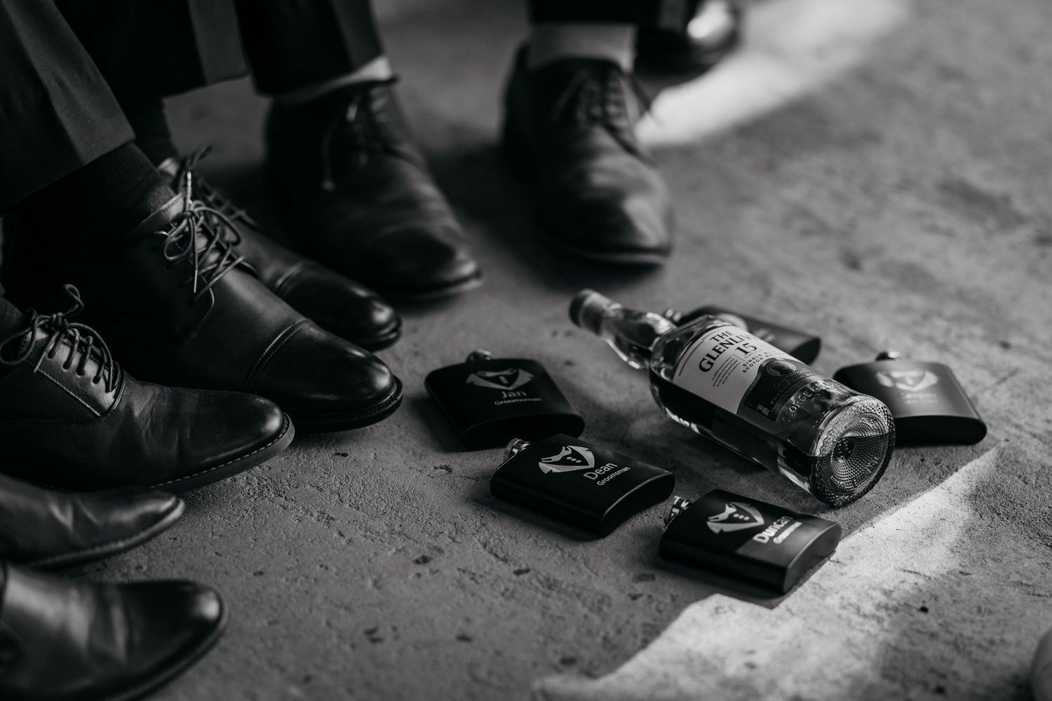 Black and white photo of dress shoes, whiskey bottle, and accessories on concrete floor at a wedding.