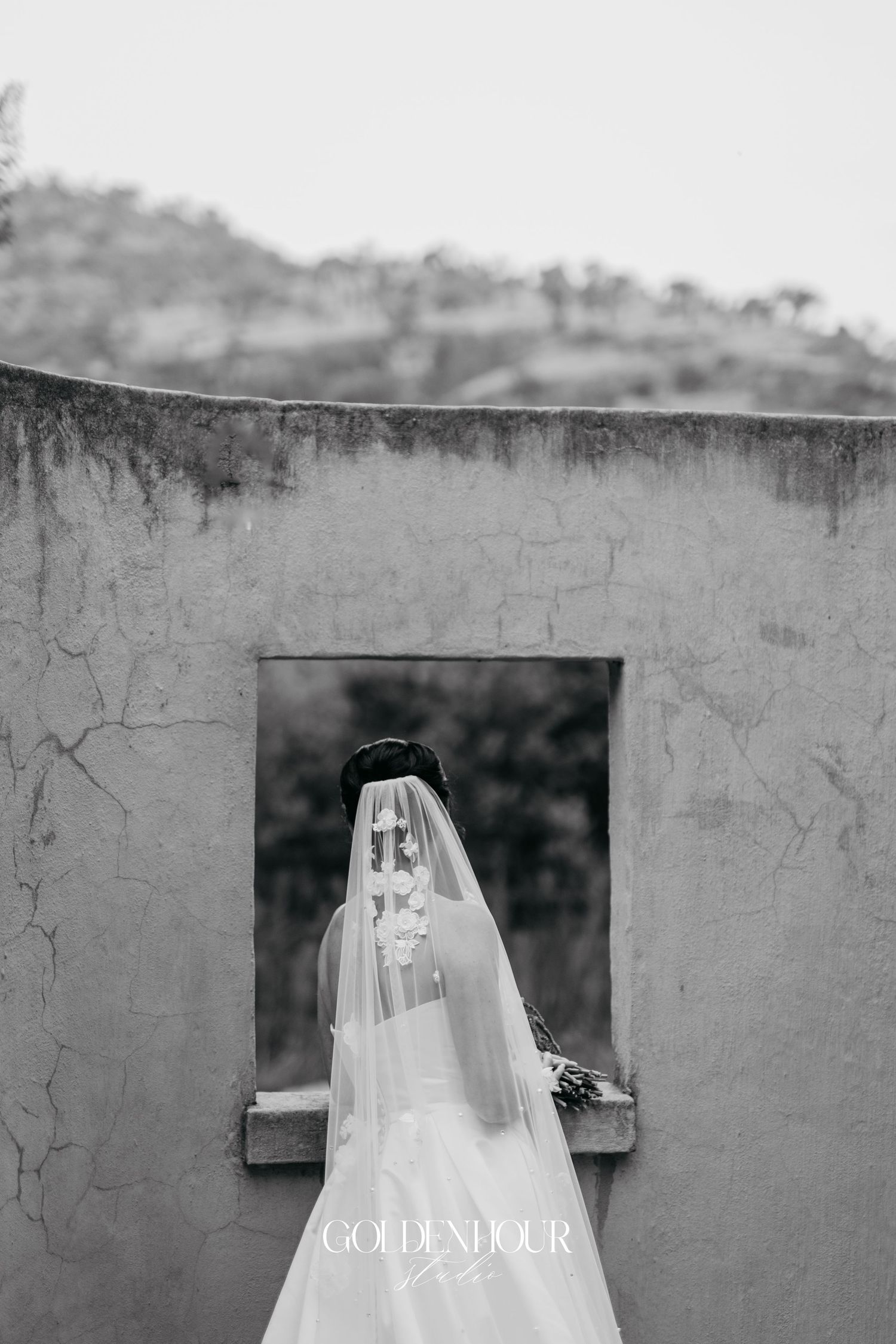 Black and white artistic photo of a veiled bride standing at a rustic stone window overlooking scenic hills.