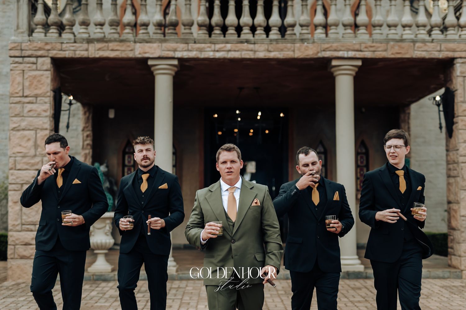 Groom in olive suit stands with groomsmen in black suits and gold ties, smoking cigars outside a elegant venue.