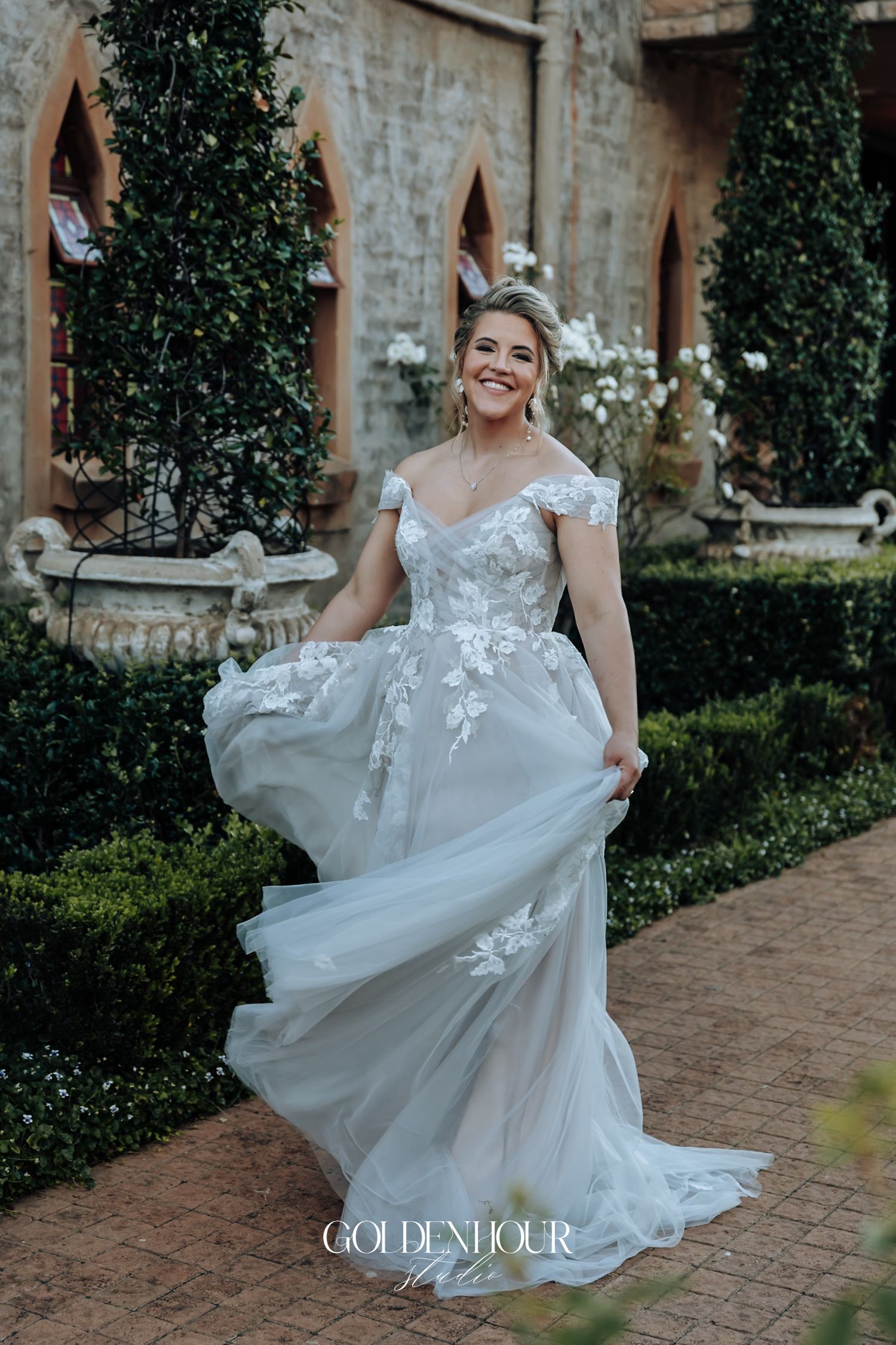 Bride twirling in a floral lace off-shoulder wedding gown in a lush garden courtyard setting.