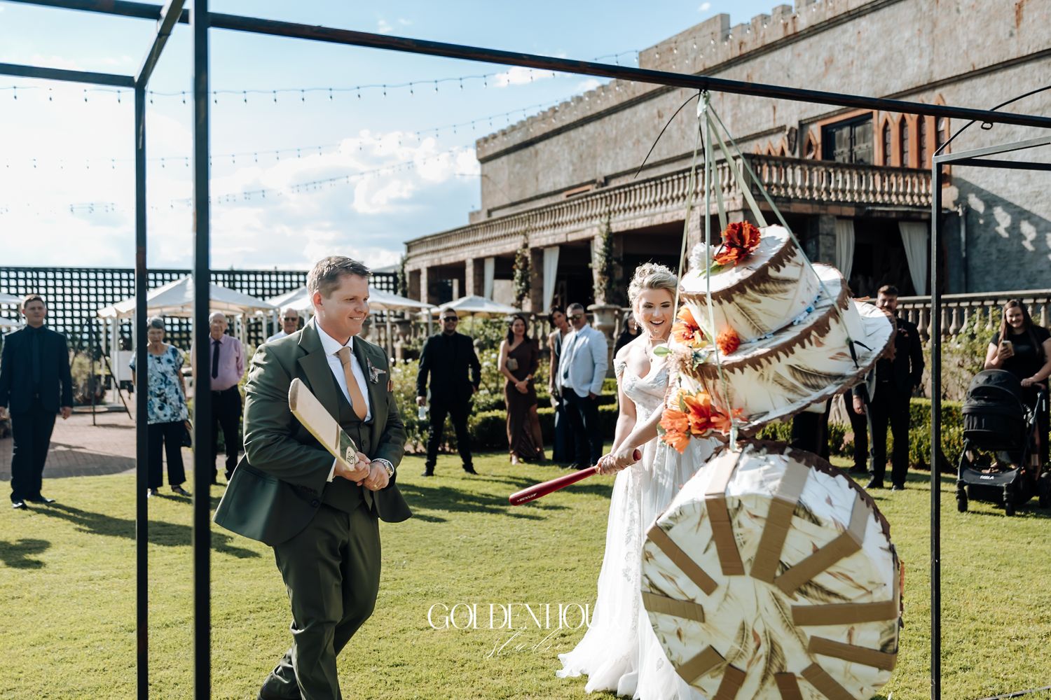 Bride and groom breaking a colorful pinata during an outdoor wedding ceremony on a green lawn.