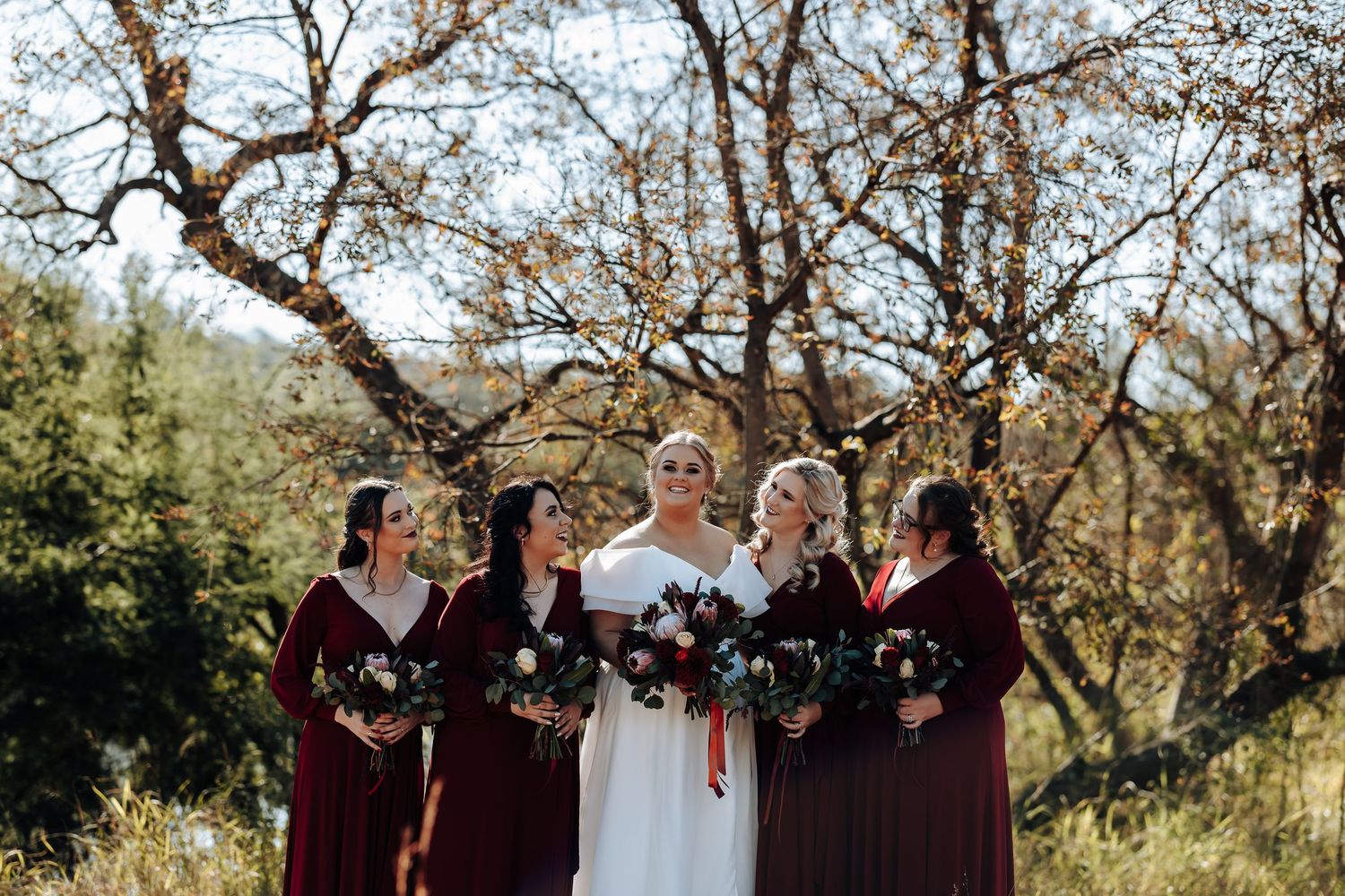 Bride in white gown with four bridesmaids in burgundy dresses holding bouquets outdoors under bare trees.
