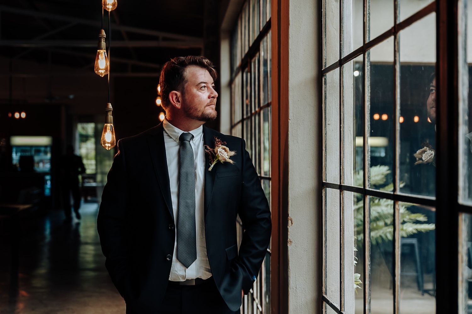 A person in a dark suit stands in a dimly lit hallway with industrial pendant lights and large windows.