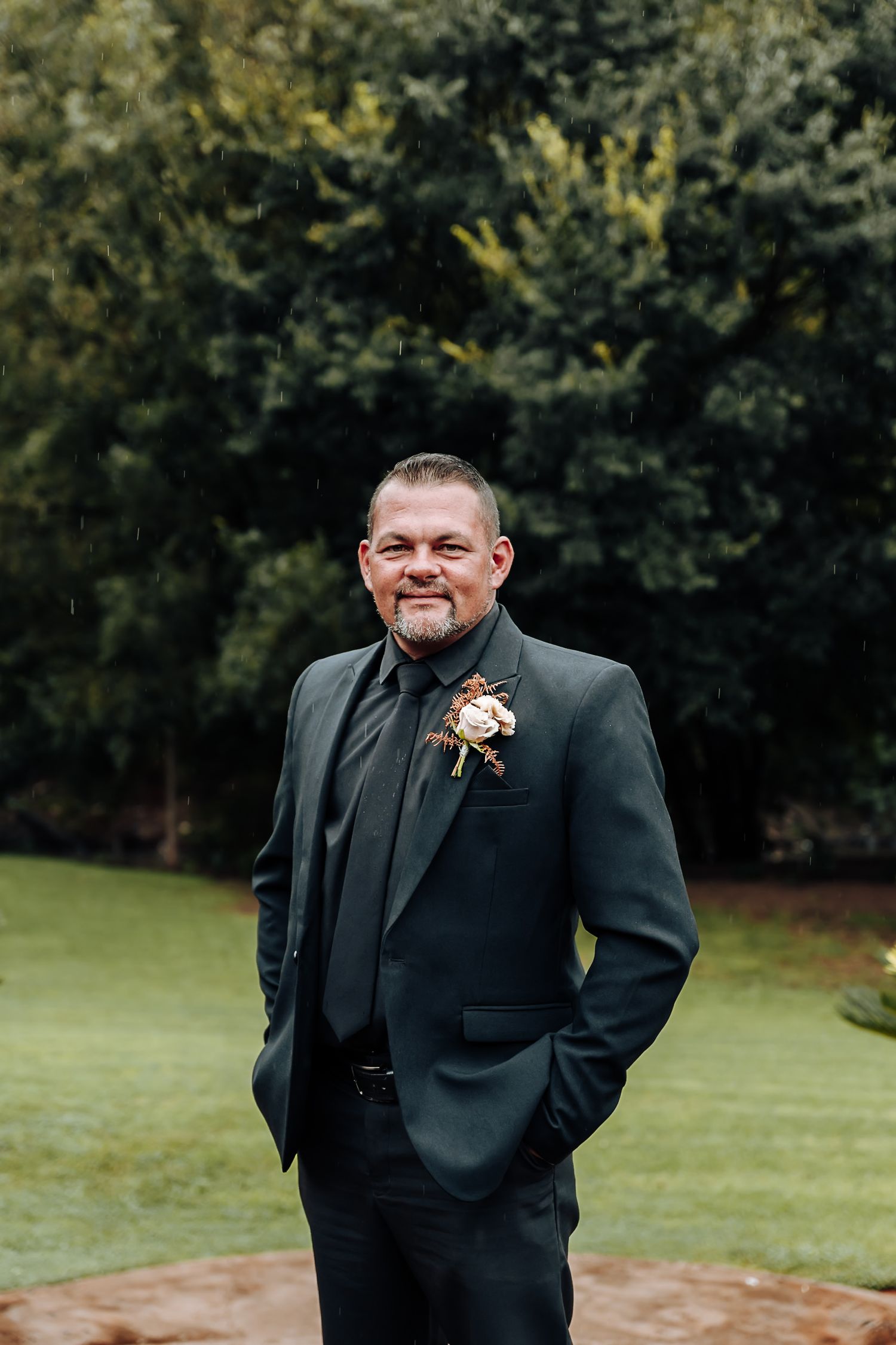 A person in formal black attire smiles warmly while standing outdoors in a natural setting with trees in background.