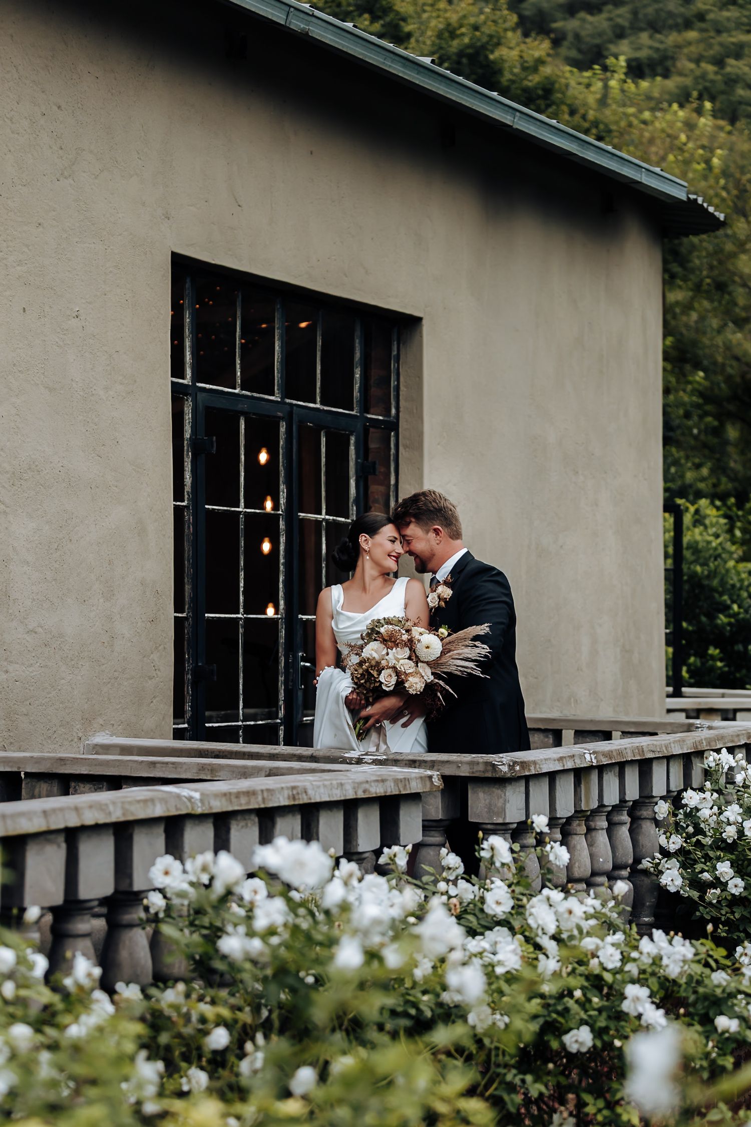 A romantic wedding couple embraces on a balcony surrounded by white roses at an elegant venue with rustic walls.
