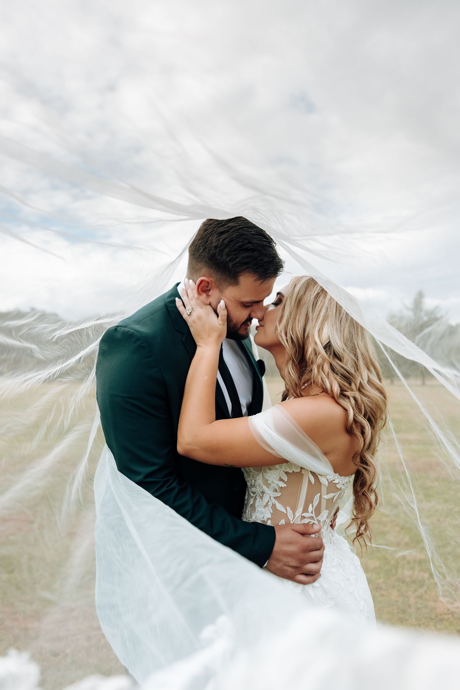 A romantic wedding couple embraces and kisses while a flowing veil creates an ethereal effect in the mountains.