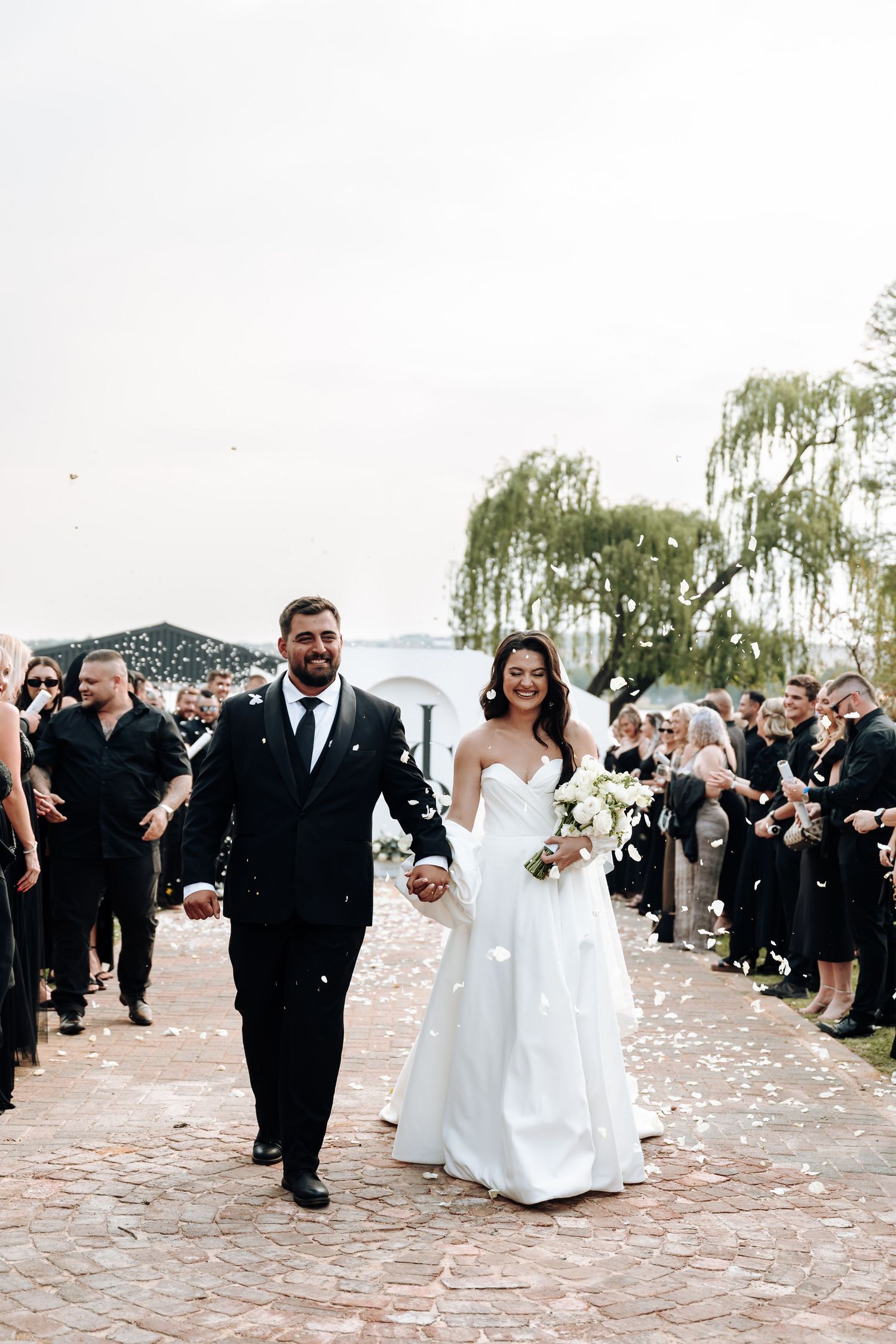 Wedding party exits ceremony through a line of guests throwing flower petals on a brick pathway.