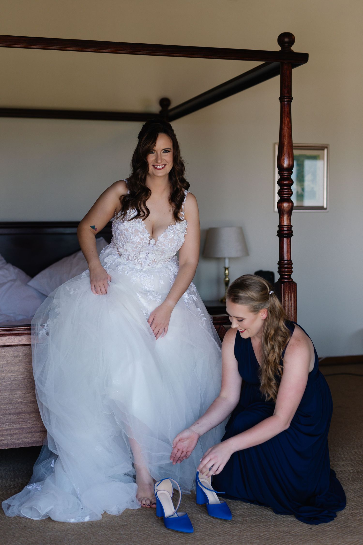 A bride in a white lace ballgown gets help putting on bright blue heels from an attendant in a navy dress.