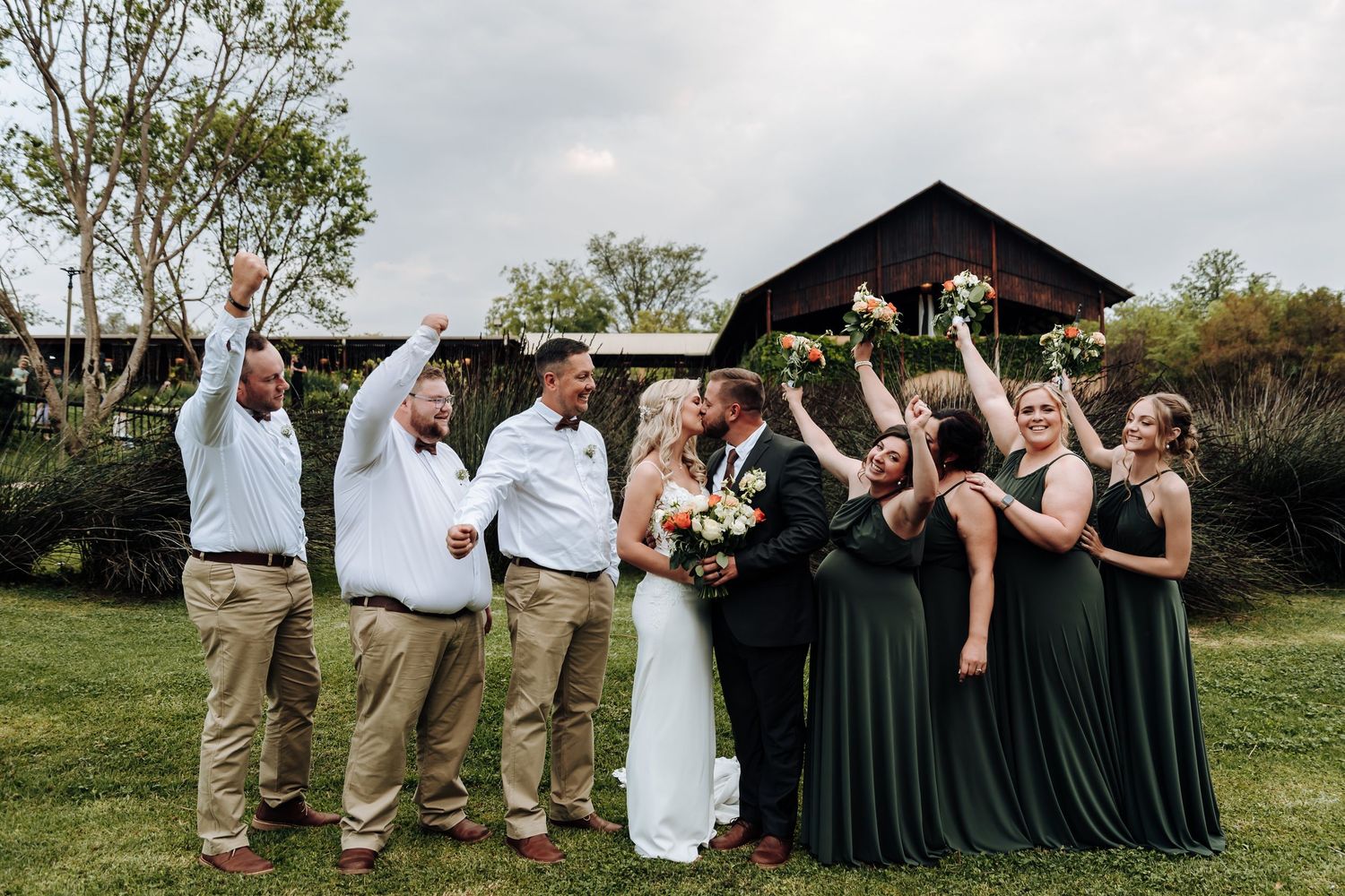 A wedding party in dark green dresses and khaki suits poses for photos in a scenic outdoor setting.