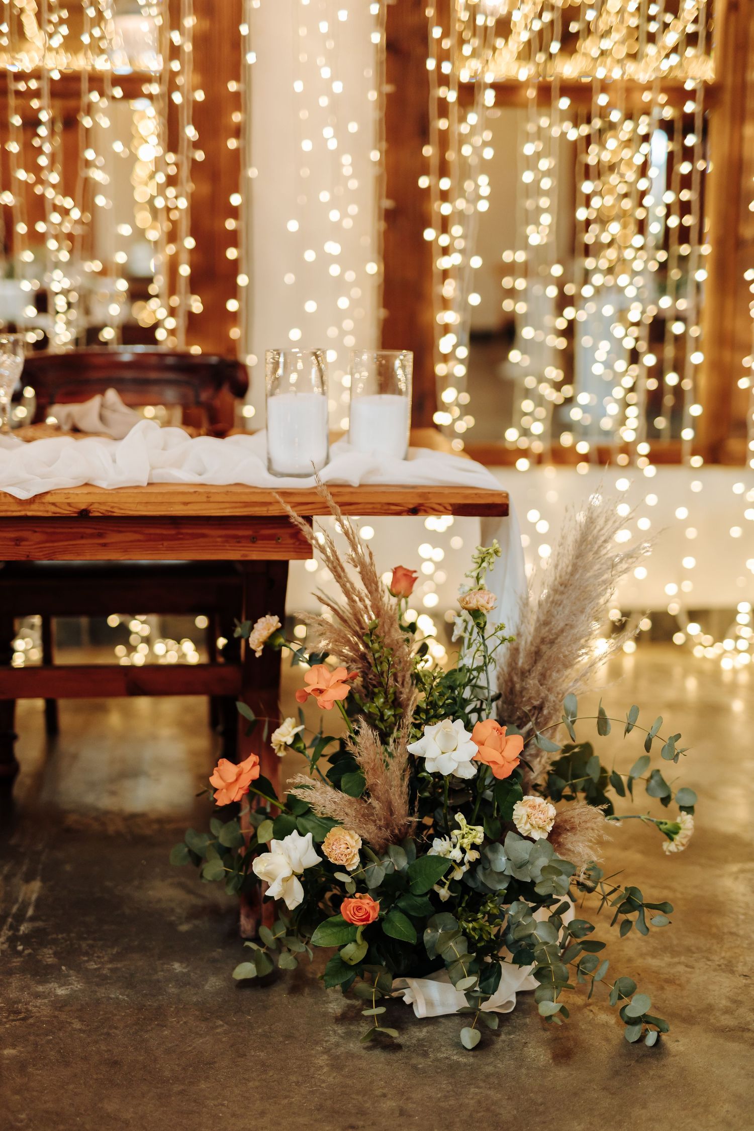 Rustic wedding reception decor with twinkle lights and floral arrangement featuring peach roses and pampas grass.