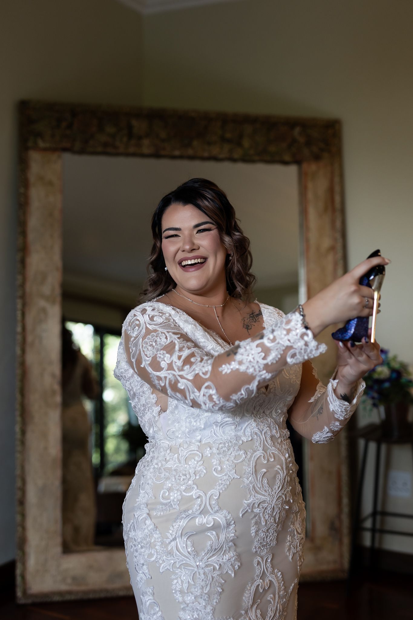 A joyful bride laughs while holding a bottle in her detailed lace wedding gown.