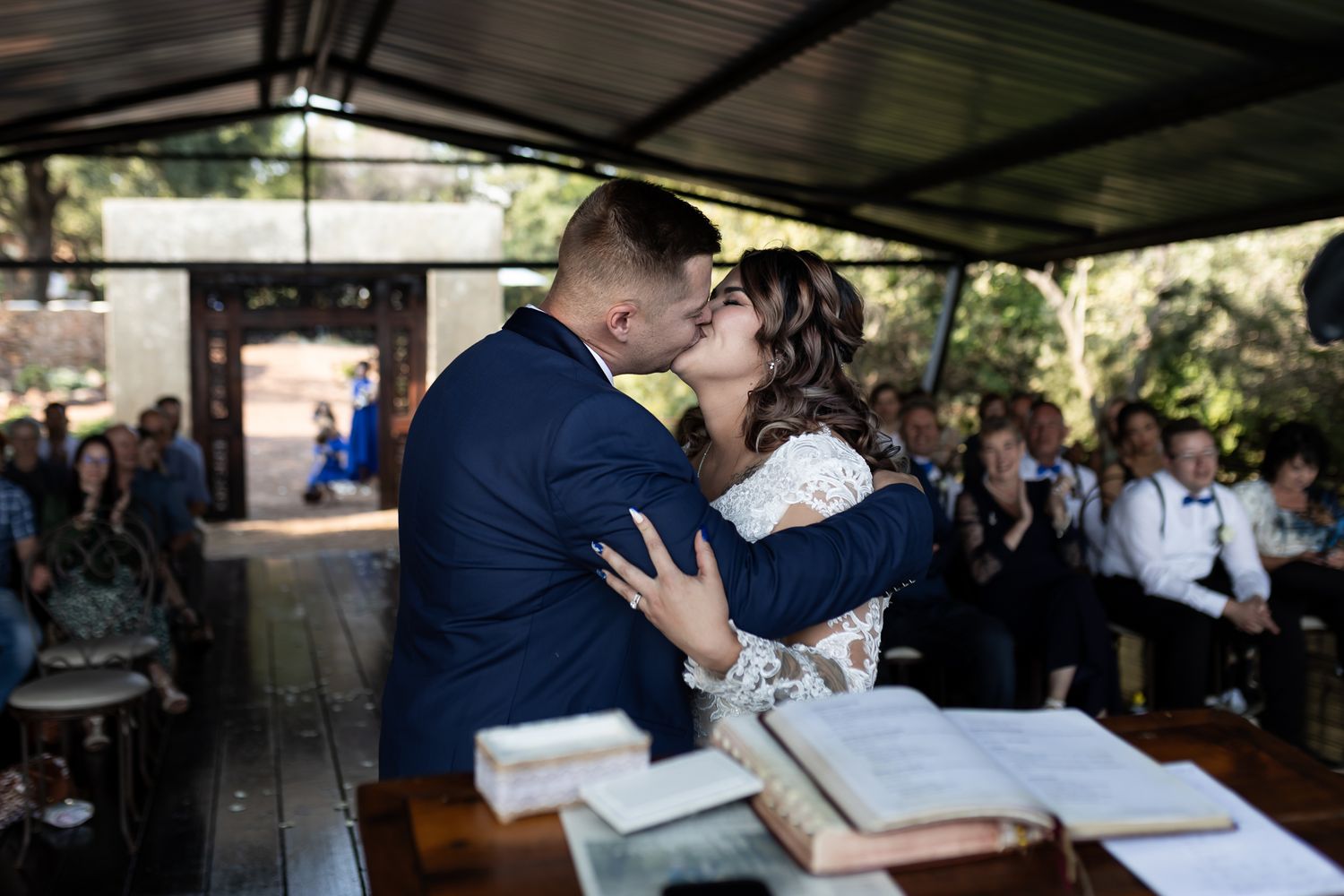 Wedding couple embraces during ceremony at rustic outdoor venue with guests in background.