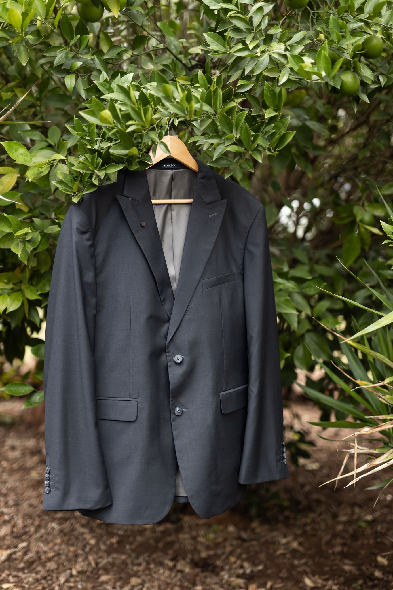 Navy blue suit jacket hanging on wooden hanger against green foliage background.