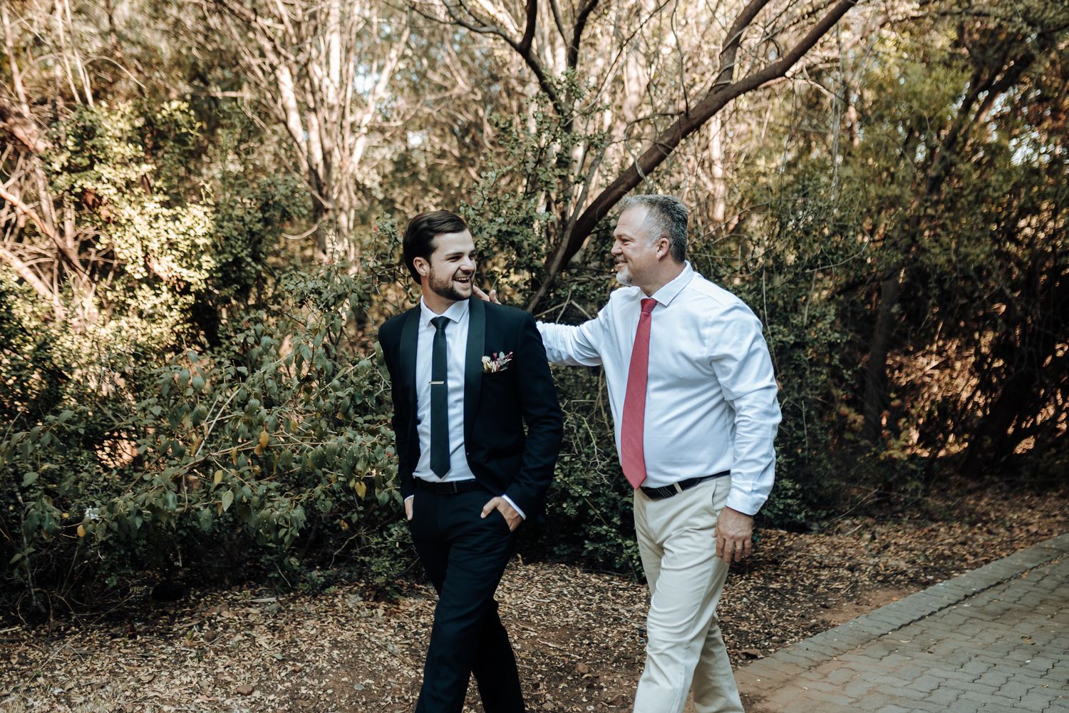 Two people in formal attire walking and conversing on a garden path surrounded by trees.