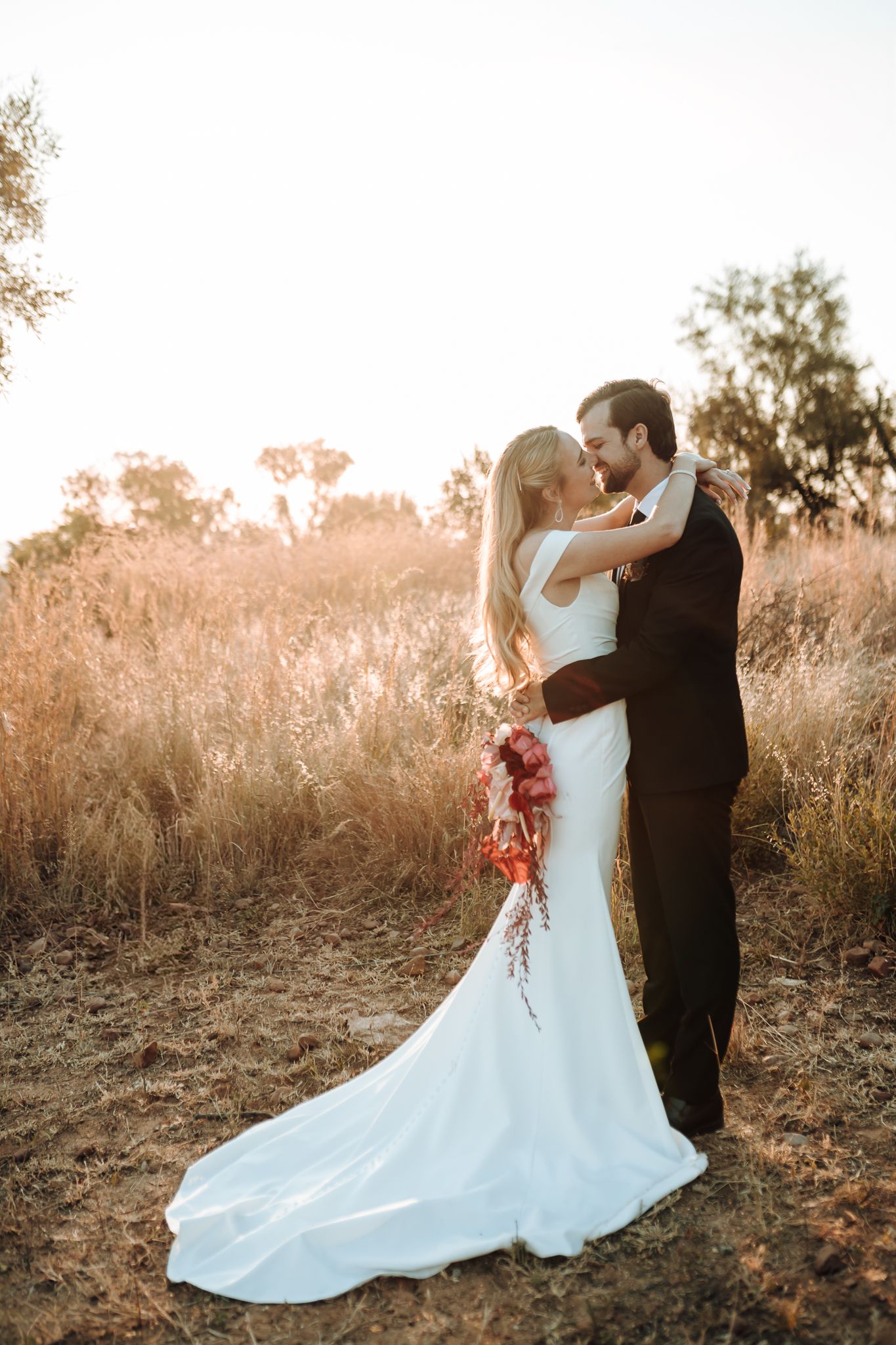 Newlyweds share an intimate moment in a sun-drenched field at golden hour with the bride's flowing white dress.