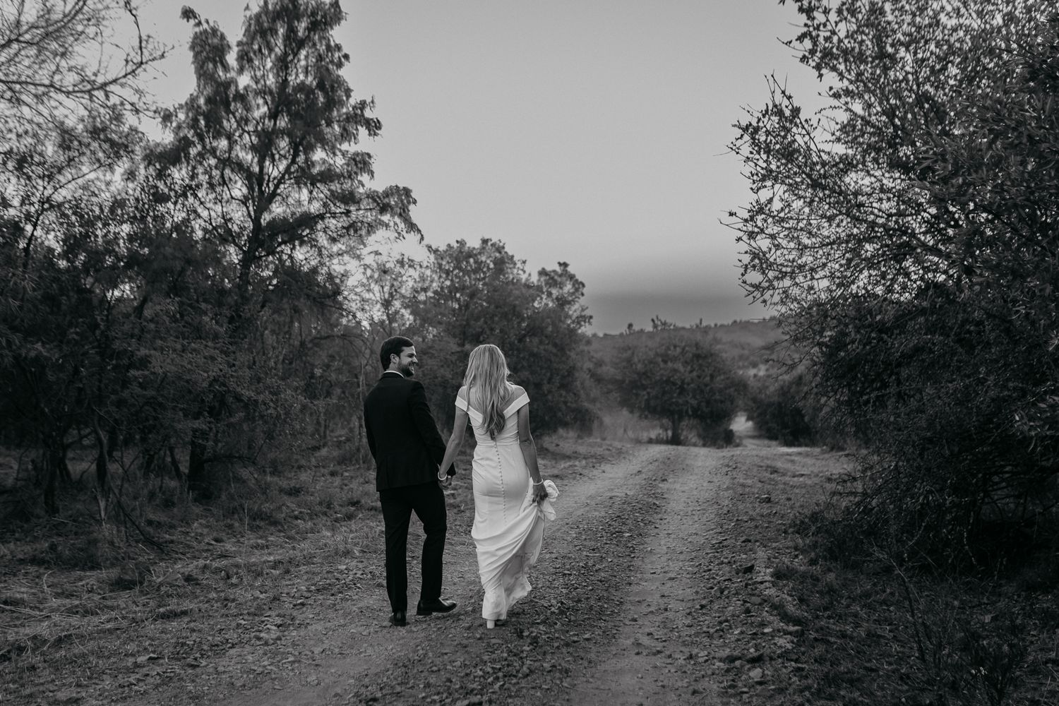 A romantic black and white photo of a couple walking down a dusty country road surrounded by trees at dusk.