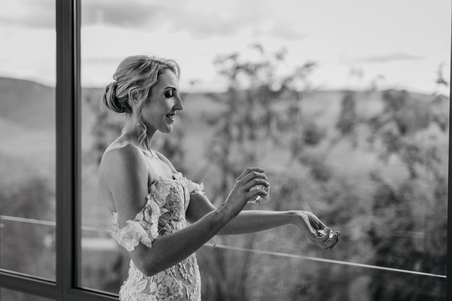 Candid black and white photo of bride in lace gown standing by scenic window view.