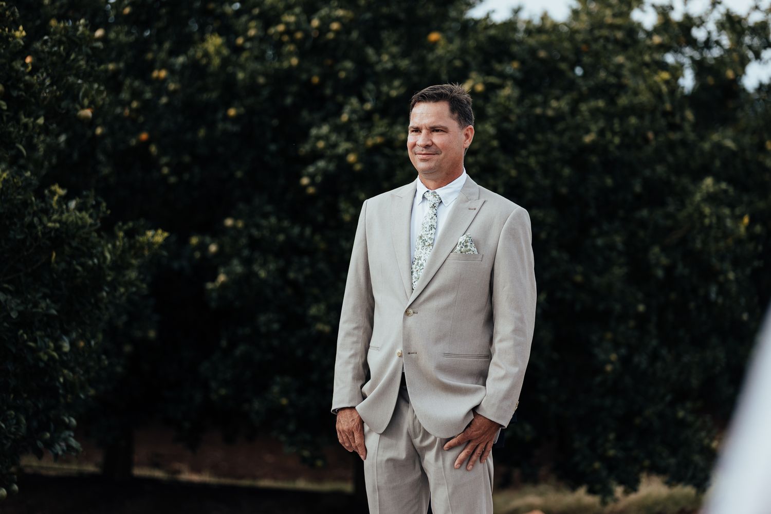 Groom in light beige suit stands ready during outdoor orchard wedding ceremony.