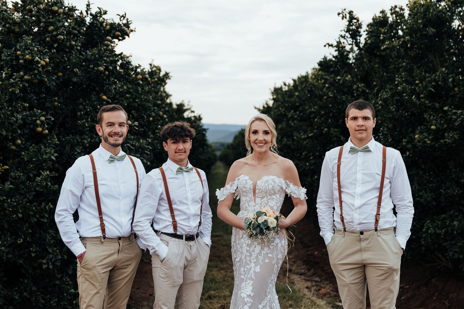Wedding party in casual attire with suspenders and light colors stand together in an orchard.