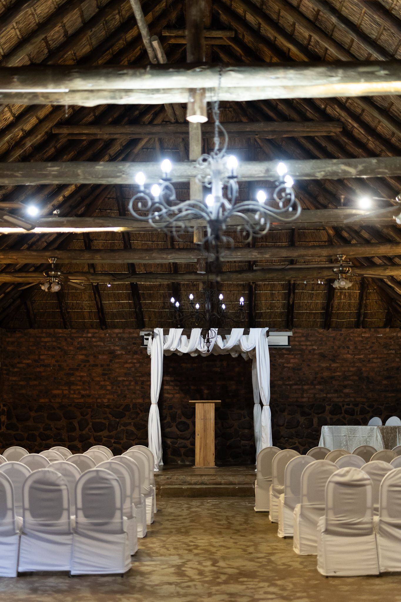 An elegant rustic wedding ceremony setup with white chairs under a wooden beam ceiling with chandeliers.