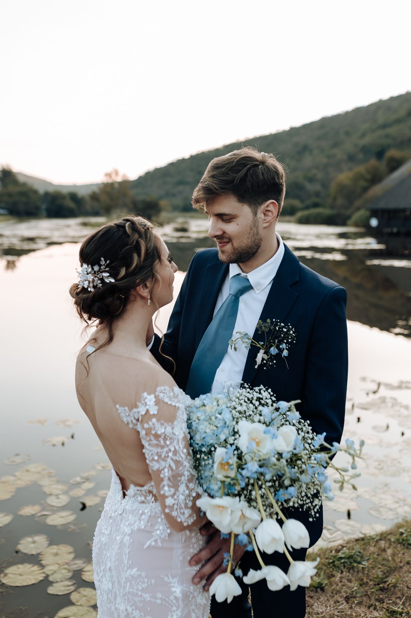 A romantic moment captured between newlyweds beside a lake with lily pads, featuring a blue and white bouquet.