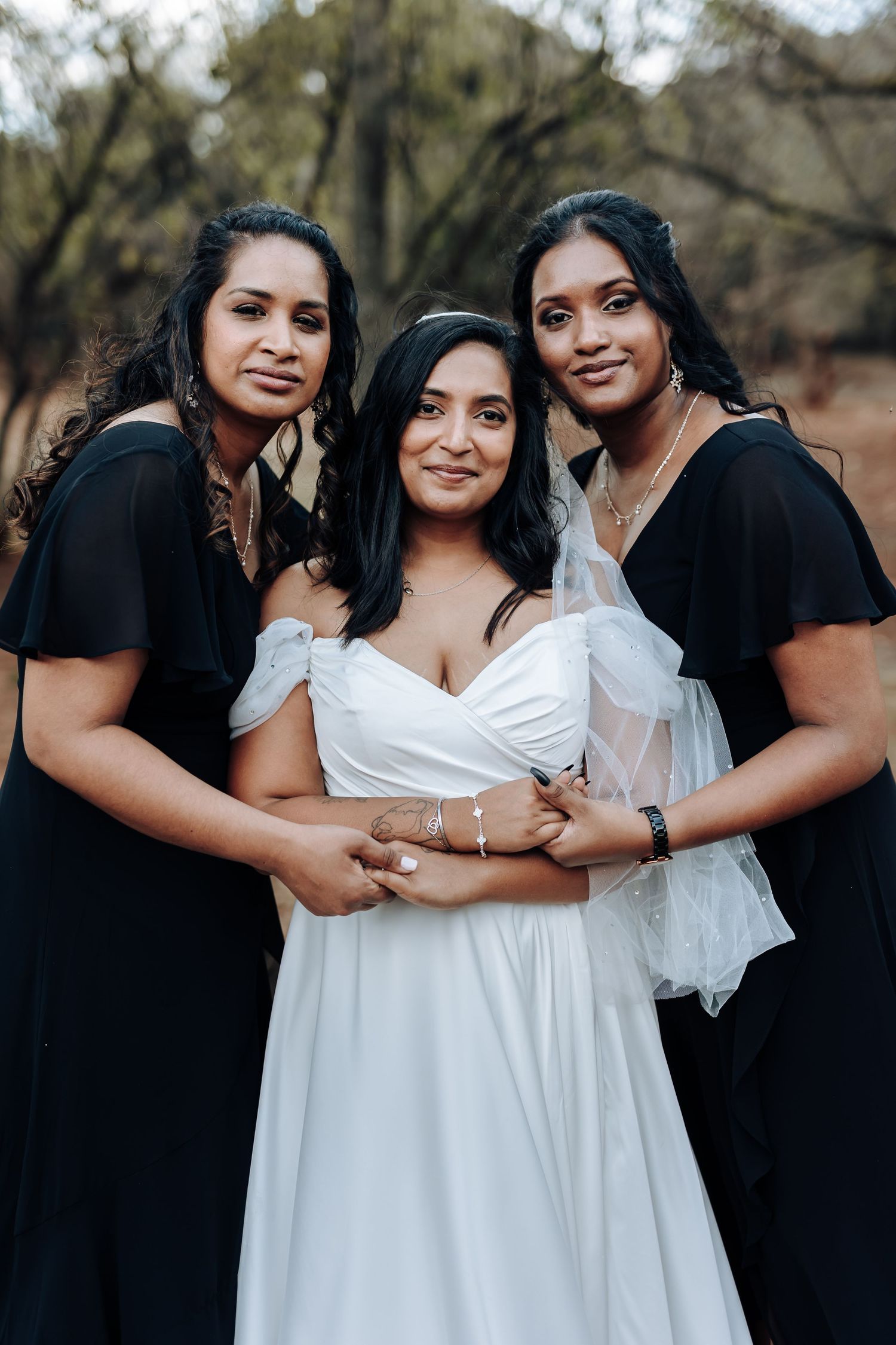 Three women embrace while wearing coordinated formal dresses outdoors.