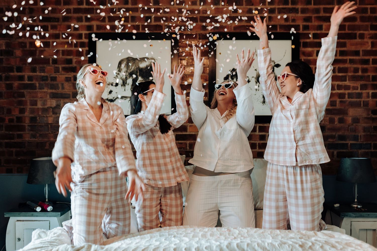 A celebratory group in matching pajamas throws confetti while jumping on a bed against a brick wall.