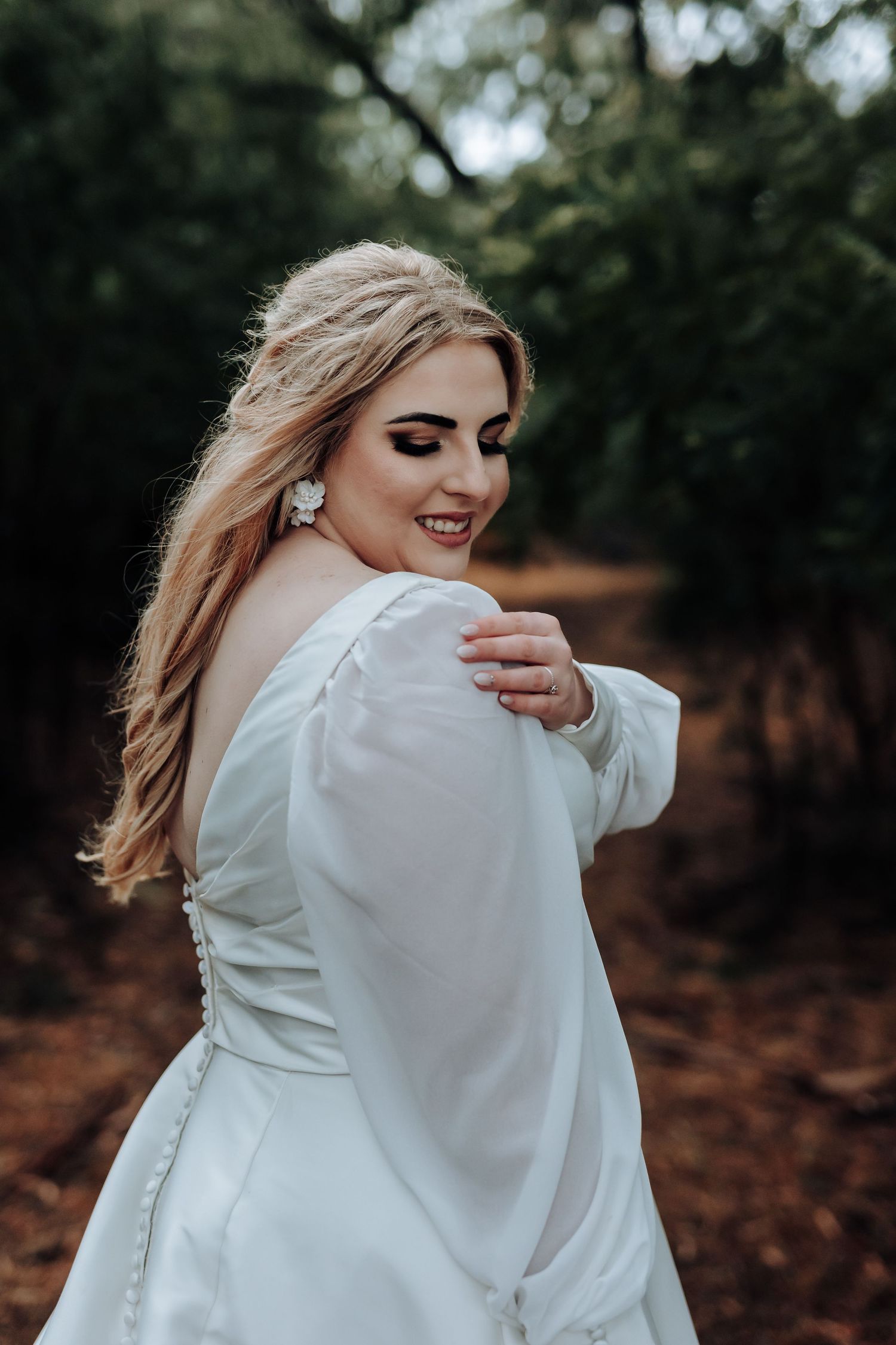 Bride in classic white gown smiles over shoulder in moody forest setting.